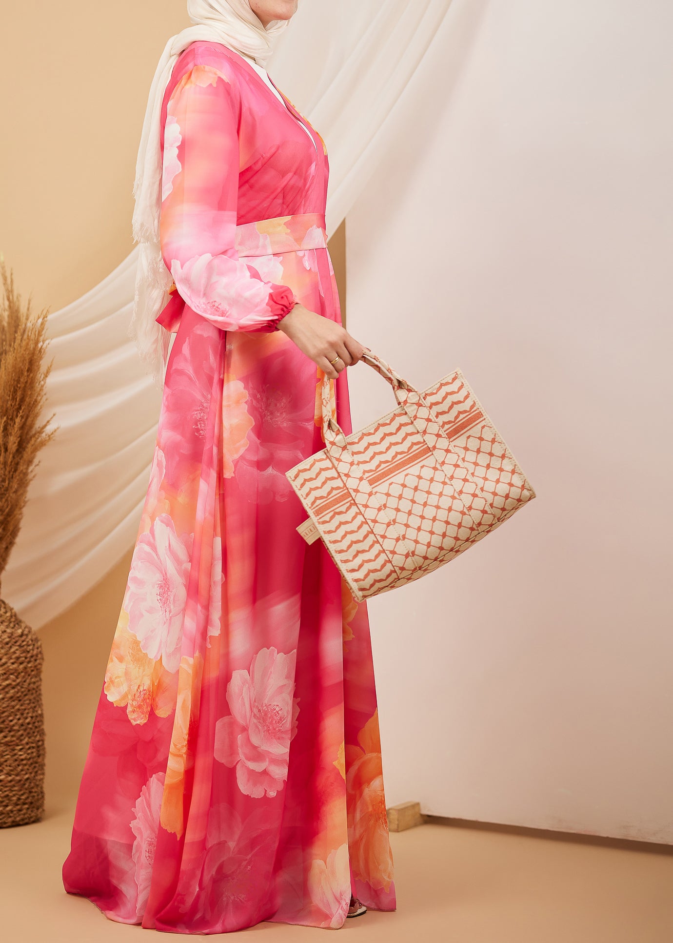 Side view of a model wearing a coral floral chiffon maxi dress, holding a woven bag