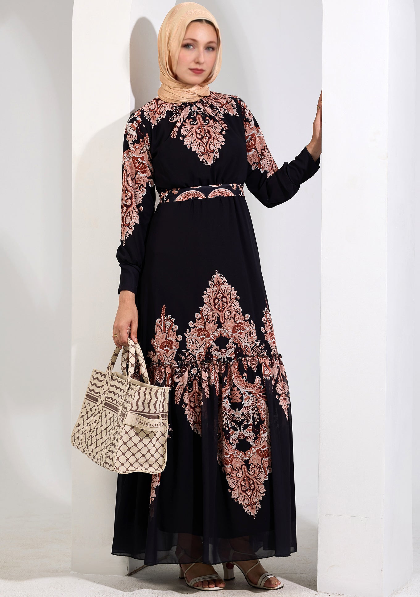 Woman holding a beige patterned handbag, wearing a black chiffon maxi dress with peach floral print