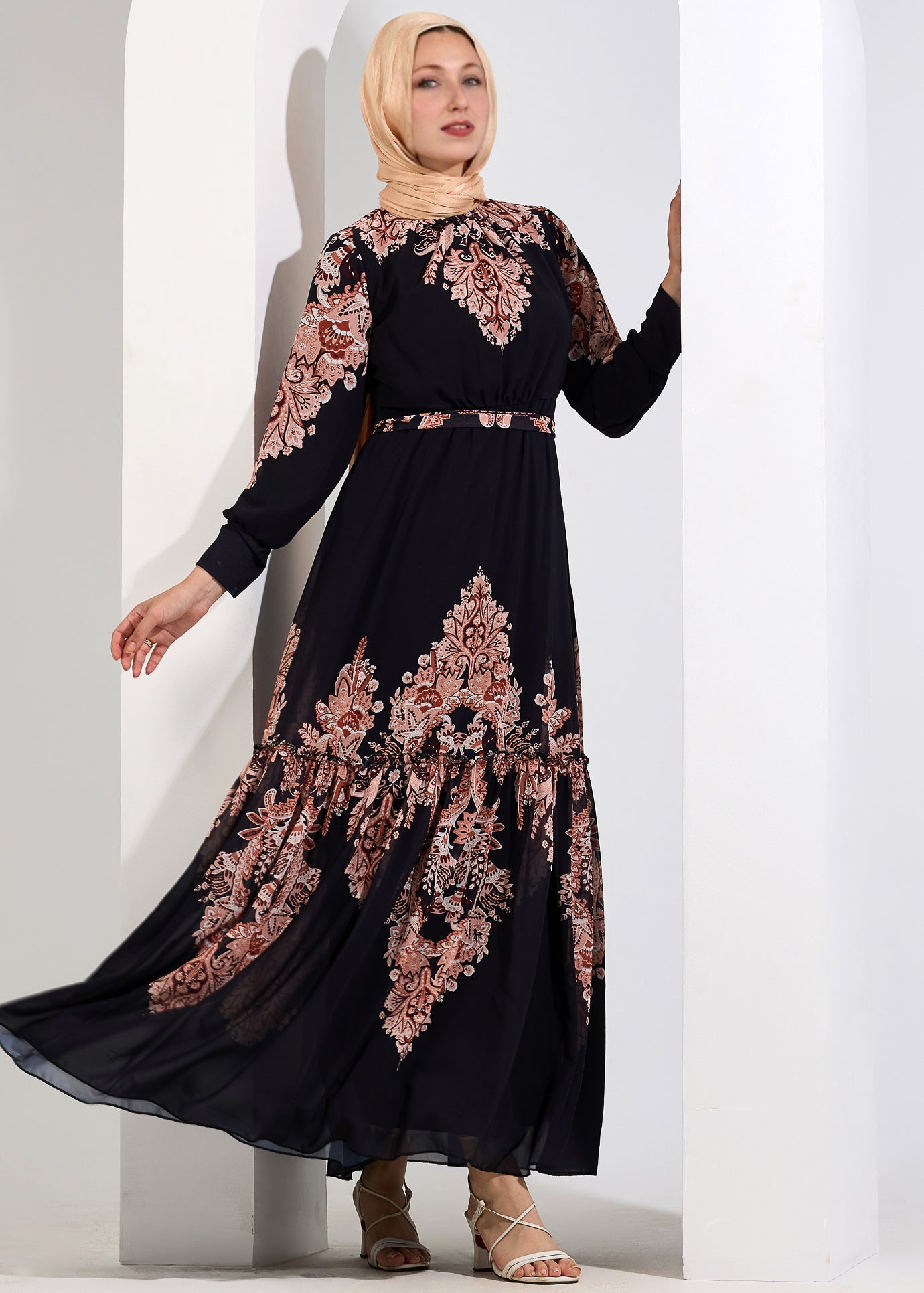 Alt Text: Woman standing in a black chiffon maxi dress with peach floral print, hands gently clasped.