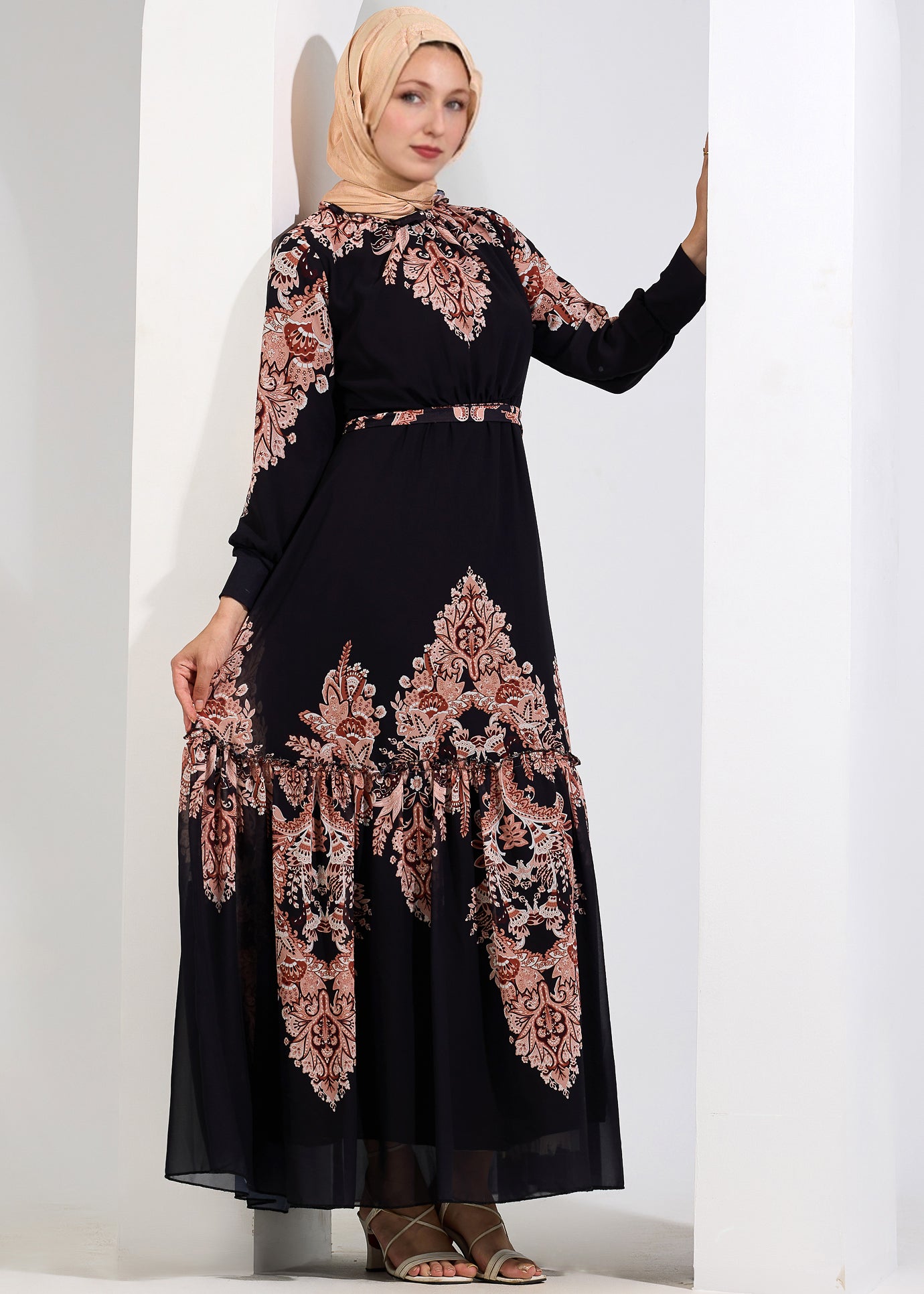 Woman posing in a black chiffon maxi dress with peach floral print, holding onto a white column