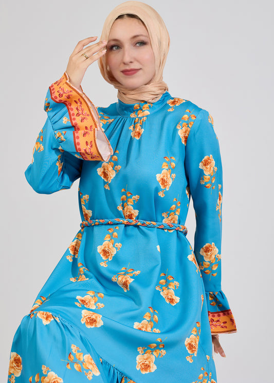 Woman wearing a turquoise floral maxi dress with ruffled sleeves, posing with one hand near her face